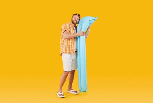 Smiling man in summer outfit is hugging inflatable pool mattress on yellow background. Full length of cheerful Caucasian man in casual summer clothes and sunglasses hugging big blue air mattress.