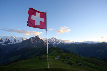 Sunrise over alpine peaks with a flag proudly displayed on a grassy ridge.