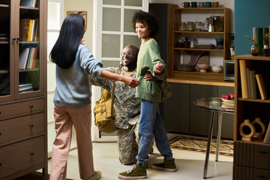 American middle aged man in military uniform kneeling and embracing smiling American woman and child in home setting, family members greeting returning soldier