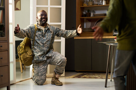 Black middle aged man in military uniform kneeling with arms open, smiling and looking toward young child entering home, family reunion scene in domestic setting - Powered by Adobe