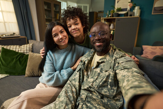 Portrait of Black middle aged man in military uniform sitting on sofa with Caucasian young adult woman and multiethnic child smiling