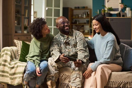 Black middle aged man in military uniform sitting on couch holding digital tablet, smiling with young adult Black woman and young adult Black man, family spending time together at home