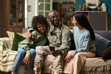 Black middle aged man in military uniform sitting on couch with arm around smiling Black child and smiling Caucasian woman, holding smartphone and spending time together at home