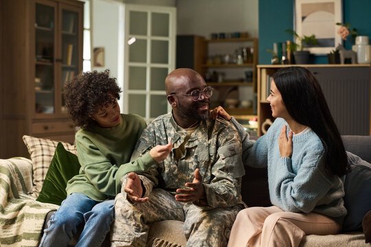 Black middle aged man in military uniform sitting on couch smiling and talking with two young adult women of different ethnicities at home, family members interacting closely - Powered by Adobe