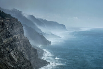 Fototapeta premium Misty fog rolls over rugged coastal cliffs and forested hillsides overlooking the ocean