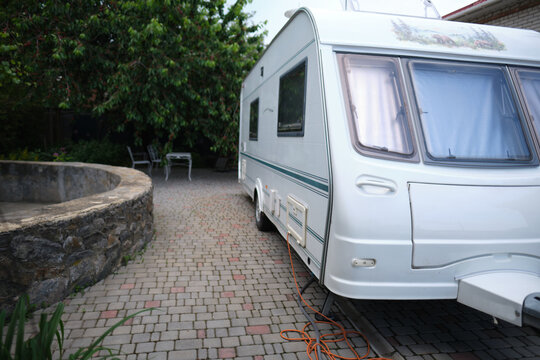 A white camper trailer is parked on a paved backyard near a brick house and cherry tree. RV travel content, camping promotions, suburban lifestyle visuals, or articles about alternative housing