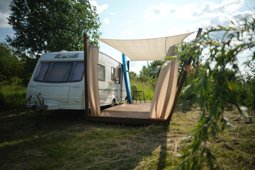 A camper van with a wooden terrace and fabric canopy is parked in the middle of tall grass and trees. This peaceful scene evokes a sense of freedom, glamping, and off-grid living.
