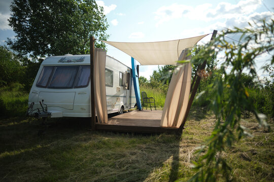 A camper van with a wooden terrace and fabric canopy is parked in the middle of tall grass and trees. This peaceful scene evokes a sense of freedom, glamping, and off-grid living.