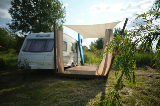 A camper van with a wooden terrace and fabric canopy is parked in the middle of tall grass and trees. This peaceful scene evokes a sense of freedom, glamping, and off-grid living.