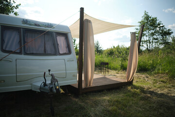 A camper van with a wooden terrace and fabric canopy is parked in the middle of tall grass and trees. This peaceful scene evokes a sense of freedom, glamping, and off-grid living.