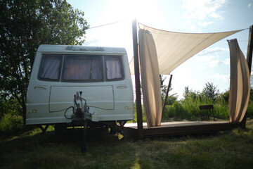 A camper van with a wooden terrace and fabric canopy is parked in the middle of tall grass and trees. This peaceful scene evokes a sense of freedom, glamping, and off-grid living.