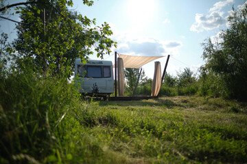 A camper van with a wooden terrace and fabric canopy is parked in the middle of tall grass and trees. This peaceful scene evokes a sense of freedom, glamping, and off-grid living.