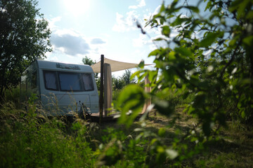 A camper van with a wooden terrace and fabric canopy is parked in the middle of tall grass and trees. This peaceful scene evokes a sense of freedom, glamping, and off-grid living.