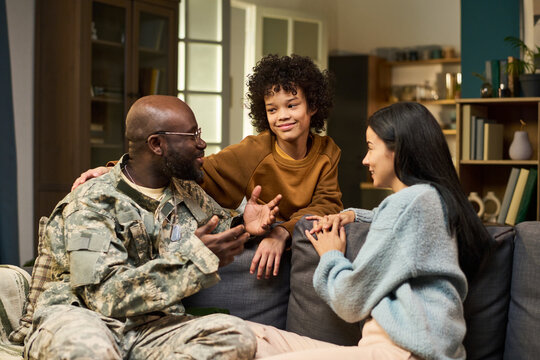 Black middle aged man in military uniform sitting on couch talking with smiling Asian woman and teenager, family members interacting together in home living room