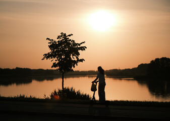 Silhouette of a girl riding a scooter past a lake at sunset, reflected on the surface of the water....