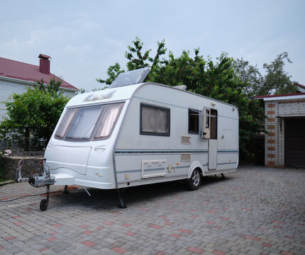 A white camper trailer is parked on a paved backyard near a brick house and cherry tree. RV travel content, camping promotions, suburban lifestyle visuals, or articles about alternative housing