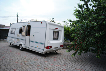 A white camper trailer is parked on a paved backyard near a brick house and cherry tree. RV travel content, camping promotions, suburban lifestyle visuals, or articles about alternative housing