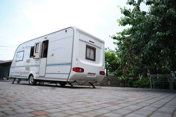 A white camper trailer is parked on a paved backyard near a brick house and cherry tree. RV travel content, camping promotions, suburban lifestyle visuals, or articles about alternative housing