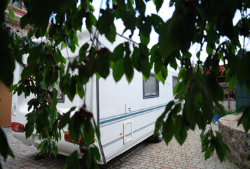 A white camper trailer is parked on a paved backyard near a brick house and cherry tree. RV travel content, camping promotions, suburban lifestyle visuals, or articles about alternative housing