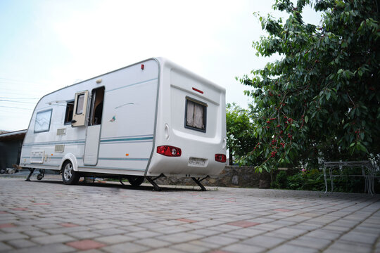 A white camper trailer is parked on a paved backyard near a brick house and cherry tree. RV travel content, camping promotions, suburban lifestyle visuals, or articles about alternative housing
