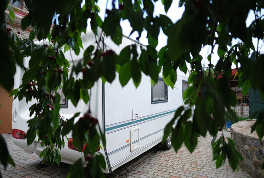A white camper trailer is parked on a paved backyard near a brick house and cherry tree. RV travel content, camping promotions, suburban lifestyle visuals, or articles about alternative housing