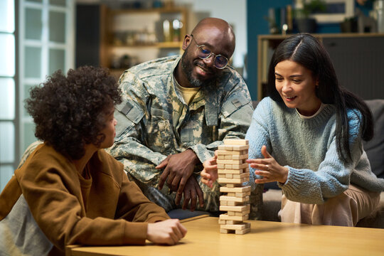 Caucasian woman and child playing wooden block game with smiling Black middle aged man in military uniform watching at home, family spending quality time together in living room - Powered by Adobe