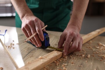Man measuring plank with tape and pencil at wooden table indoors, closeup