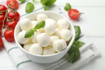 Tasty mozzarella cheese balls in bowl, basil and tomatoes on white wooden table, closeup