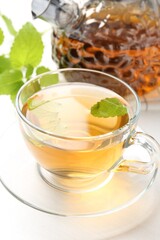Aromatic lemon balm tea in glass cup, teapot and fresh leaves on white table, closeup