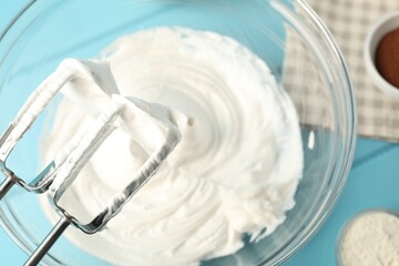 Bowl with whipped cream, hand mixer and ingredients on light blue wooden table, flat lay