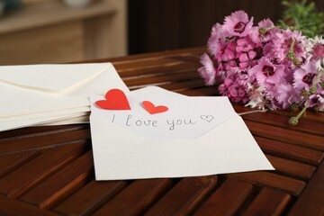 Card with phrase I Love You, letters, red hearts and flowers on wooden table indoors, closeup
