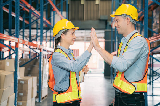 Confidence caucasian man, woman staff worker wear safety helmets, vest high five teamwork collaborate, cooperation in logistics inventory operation. show success,efficiency modern warehouse management