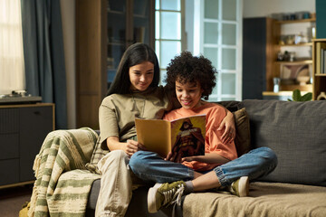 Caucasian young adult woman sitting on sofa embracing multiethnic teenager girl reading book...
