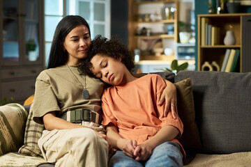 Caucasian young adult woman with military dog tags sitting on sofa embracing multiethnic teenager...
