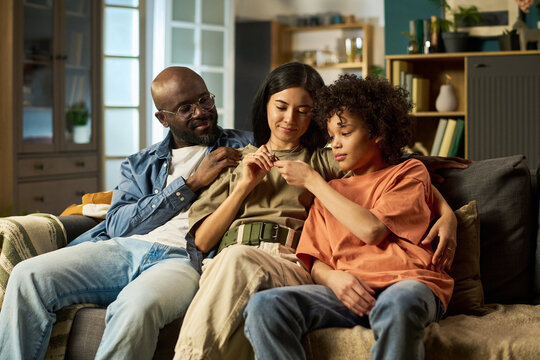 Black middle aged man in military style clothing sitting on sofa with young adult Caucasian woman and teenage Black boy, family members interacting closely at home