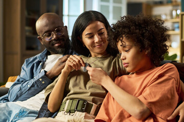 Caucasian young adult woman, Black middle aged man, and Black child sitting together on sofa,...