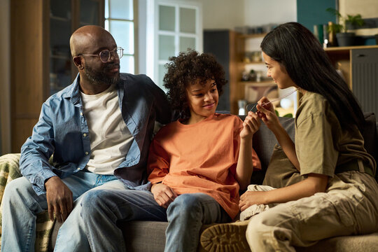 Black middle aged man sitting with two teenagers on sofa, smiling and interacting together at home, teenage girl showing necklace to teenage boy, family spending time in living room - Powered by Adobe