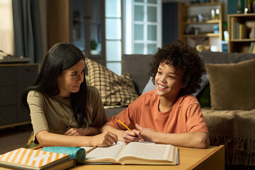 Caucasian woman helping smiling Black teenager with homework at home, both sitting at table with...