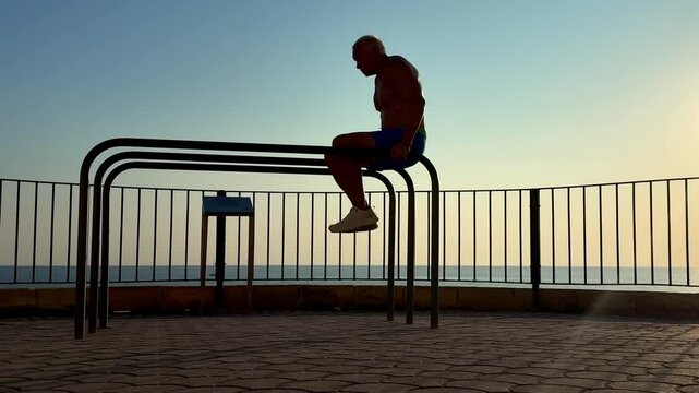 Tanned elderly man working out on parallel bars by the sea at sunrise or sunset. Silhouette, street workout, senior fitness, healthy lifestyle, inspiring active aging outdoor scene.