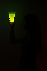 Silhouette of woman holding glowing green drink in dark setting with eerie lighting creating spooky mysterious Halloween themed party atmosphere