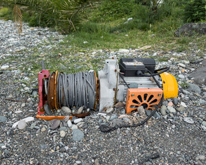 Old portable electric winch with metal cable abandoned on rocky coastal terrain. Rusty machinery surrounded by pebbles and grass. Industrial equipment outdoors