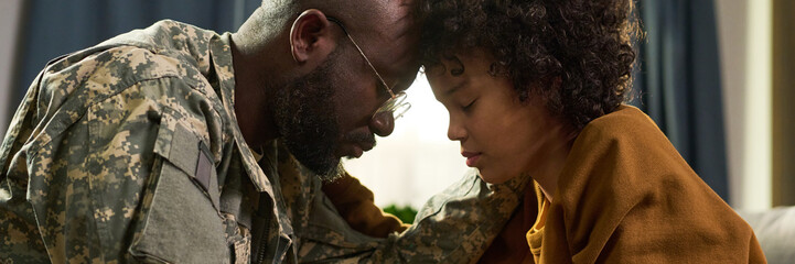 Obraz premium Black middle aged man in military uniform comforting Black young adult woman at home, both sitting closely with foreheads touching, eyes closed, sharing emotional moment together