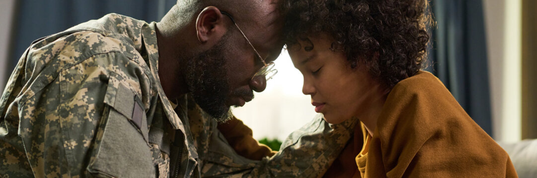 Black middle aged man in military uniform comforting Black young adult woman at home, both sitting closely with foreheads touching, eyes closed, sharing emotional moment together