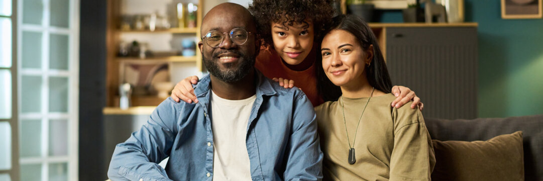 Portrait of middle aged Black man in casual clothing sitting with smiling multiethnic teenager and young adult Caucasian woman at home, all looking at camera, showing family unity - Powered by Adobe