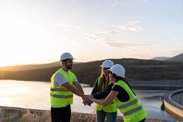 Engineers joining hands near a dam at sunset: teamwork and success in engineering projects
