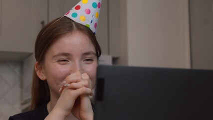 Young teen girl in colorful party hat participating in virtual birthday celebration, communicating with laptop screen, smiling and emitting cheerful emotions during online meeting