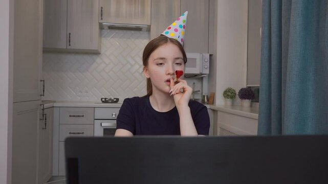 Teenage girl in party hat celebrating birthday online blowing horn during video call with piece of birthday cake and candle in front of laptop in kitchen at home.