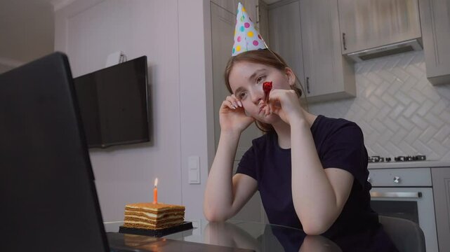 Teenage girl in party hat celebrating birthday online blowing horn during video call with piece of birthday cake and candle in front of laptop in kitchen at home. vertical video