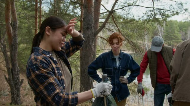 Burnette woman placing trash in plastic bag, wiping sweat from forehead and starting chatting with her friend