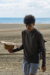 Teen boy walking on beach holding a straw hat